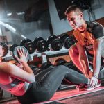 A personal trainer assists a woman doing sit-ups with a medicine ball in a gym setting.